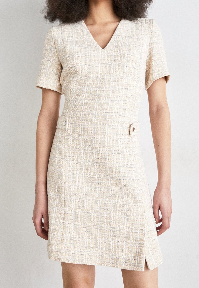 Femme portant une robe en tweed beige à manches courtes, avec un col en V, des pattes de taille boutonnées et une petite fente sur le côté gauche.