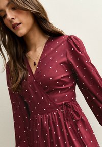 Bordeauxrotes Wickelkleid aus glattem Stoff mit weißen Tupfen, langen Puffärmeln und einer Taille mit Bindeband für eine verstellbare Passform.
