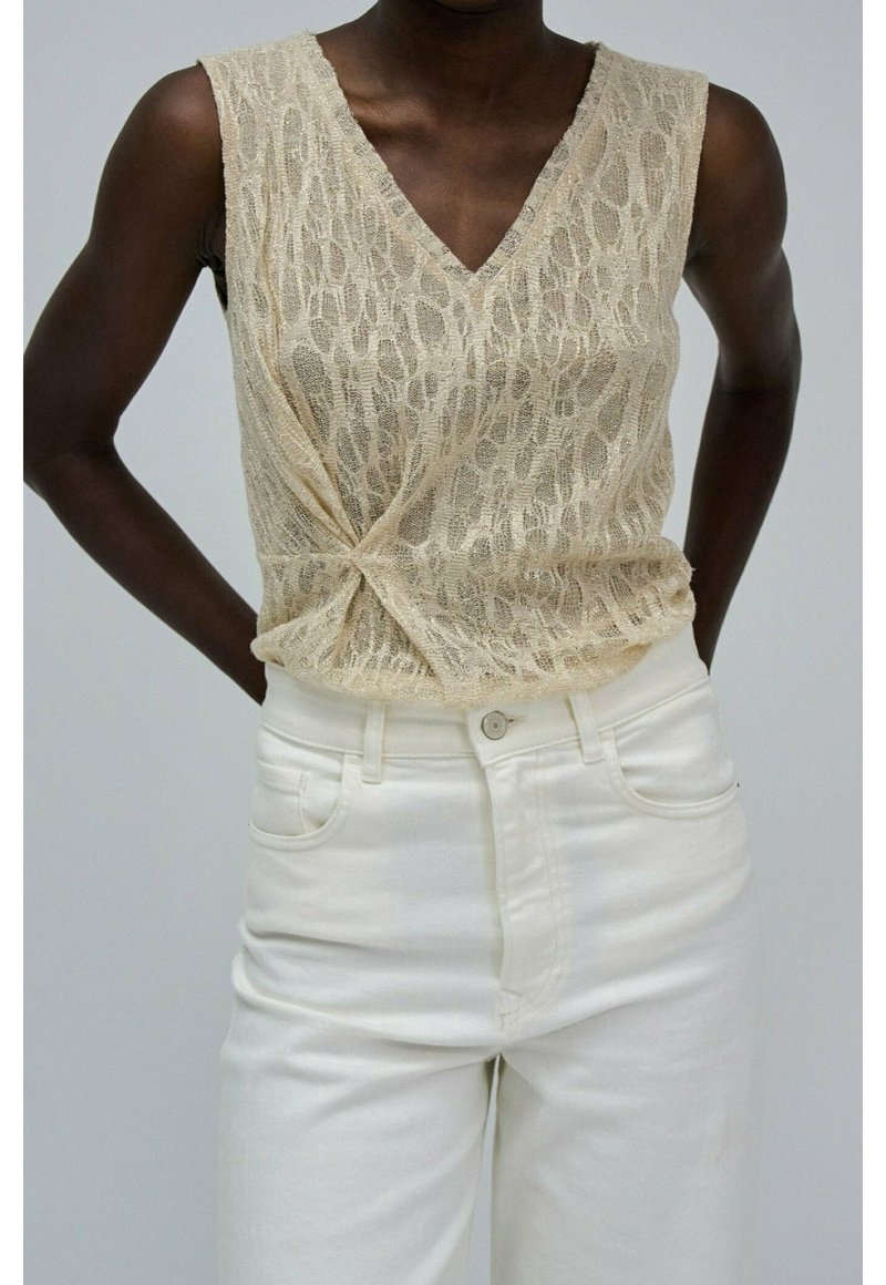 Donkerhuidige persoon in een mouwloos beige kanten top met een draai detail bij de taille en hoog-uitgesneden witte broek.
