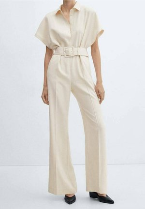 Vrouw draagt een beige short-sleeve jumpsuit met riem, met wijd uitlopende broekspijpen en zwarte puntige schoenen tegen een effen achtergrond.