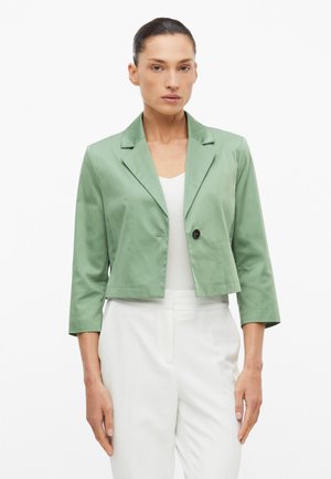 Vrouw met haar naar achteren gebonden, draagt een korte saliegroene blazer over een wit topje en witte high-waisted broek, kijkt naar voren met een neutrale uitdrukking.