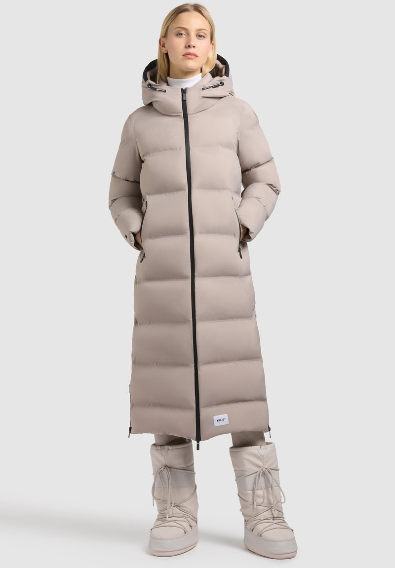Vrouw in een lange beige pufferjack met capuchon en rits, met de handen in de zakken, gecombineerd met beige winterlaarzen tegen een effen achtergrond.