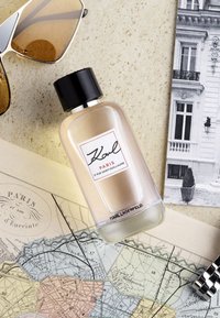 Flacon de parfum en verre transparent avec un capuchon noir, étiqueté "Karl Paris", placé sur une surface beige texturée aux côtés de lunettes de soleil et d'une carte.