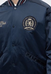 Μπουφάν bomber σε ναυτικό μπλε με κεντημένο έμβλημα που περιλαμβάνει λιοντάρι, κορώνα και ονόματα πόλεων γύρω, με κουμπιά μπροστά και ριγωτό γιακά.
