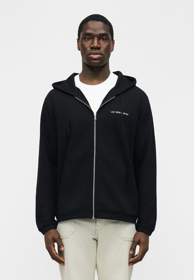 Άνδρας στέκεται προς τα εμπρός φορώντας μαύρο φούτερ με φερμουάρ και το κείμενο «Les Benjamins», λευκό πουκάμισο και ανοιχτόχρωμο παντελόνι.