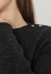 Maglione in lana grigio scuro con collo rotondo e bottoni decorativi in argento sulla spalla. La consistenza sembra morbida e accogliente.
