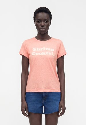 Femme aux cheveux courts et bouclés portant un t-shirt rose « Shrimp Cocktail » et un short en jean bleu, debout devant un fond blanc uni.