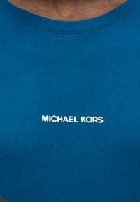 Μπλε μπλούζα με στρογγυλή λαιμόκοψη, φορεμένη από άτομο, με λευκό κείμενο "MICHAEL KORS" στο κέντρο του στήθους.