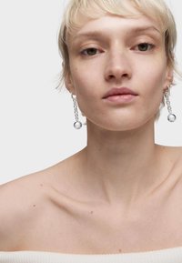 Pendientes colgantes de plata con un diseño de cadena enlazada y cuentas redondas. El modelo tiene el cabello corto y claro y lleva una blusa sin mangas.