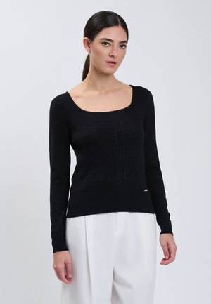 Donna con capelli scuri raccolti in una coda che indossa un maglione nero a maniche lunghe con una trama e pantaloni bianchi, in piedi davanti a uno sfondo chiaro e semplice.