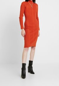 Ribbade stickade orange klänning med långa ärmar, hög hals och scalloped fåll. Buren med svarta ankelstövlar som visar en kroppsnära silhuett.