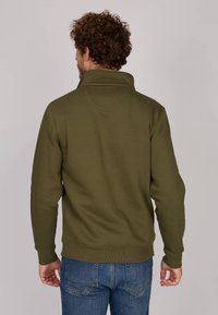 Sweatshirt vert olive avec un col haut et des poignets côtelés, d'une texture lisse et avec un motif subtil au niveau de l'ourlet. Porté avec un jean.