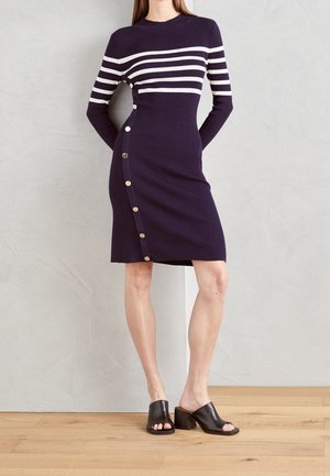 Kvinde iført en lilla ribstrikket kjole med hvide vandrette striber og metalliske knapper i siden, kombineret med sorte åbne hæle.