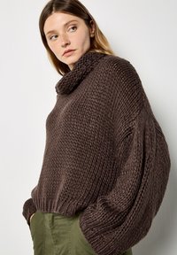 Gruby brązowy sweter z golfem, z fakturowanym wzorem i oversize'owymi rękawami. Noszony z oliwkowymi spodniami.