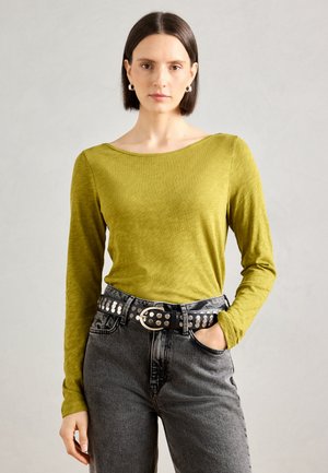 Top a maniche lunghe, verde oliva, con scollatura a barca, abbinato a jeans grigi scuri a vita alta e una cintura nera con borchie. Tessuto morbido.