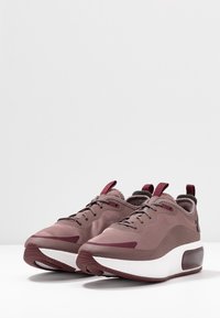 Ett par mauve träningsskor med burgundy-accenter, vita tjocka sulor och dragremmar på en vit yta mot en enkel bakgrund.