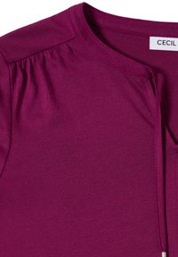 Haut fuchsia avec encolure ronde, texture douce, détails froncés aux épaules, et un lien décoratif au niveau de l'encolure. Étiquette de la marque : CECIL.