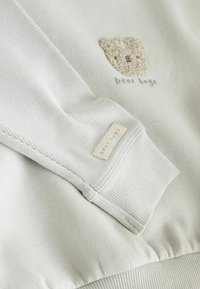 Ljusgrå sweatshirt med en texturerad björnfanspatch och broderad text "björnkramar." Inkluderar en vävd etikett på ärmslutet.