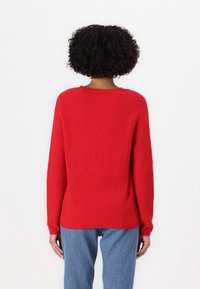 Pull rouge en tricot avec une texture côtelée, manches longues et encolure arrondie, associé à un jean en denim bleu.