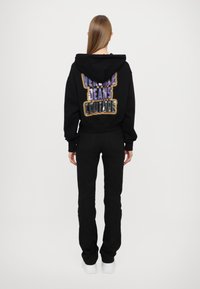 Felpa nera con cappuccio, caratterizzata da una grafica testuale multicolore "Versace Jeans Couture" sul retro. Abbinata a jeans neri e scarpe bianche.