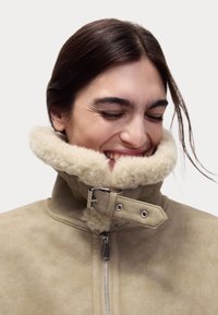 Beige schapenvachtjas met een hoge kraag, voorzien van een fluffy crèmekleurige voering en zilverkleurige hardware, inclusief een gesp en rits.