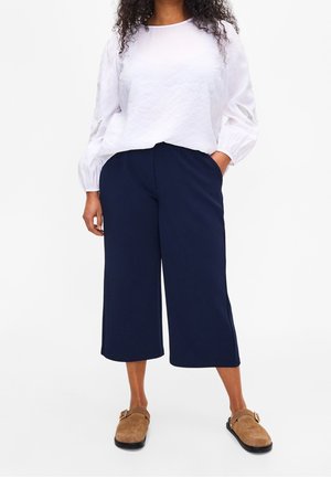 Femme portant un chemisier blanc à manches longues, un pantalon large bleu marine raccourci, et des chaussures à enfiler marron avec boucle, debout avec une main dans la poche.