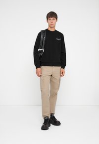 Black sweatshirt with text, beige cargo pants, and black chunky sneakers. Model holds a shiny black strap with buckles. Simple background.