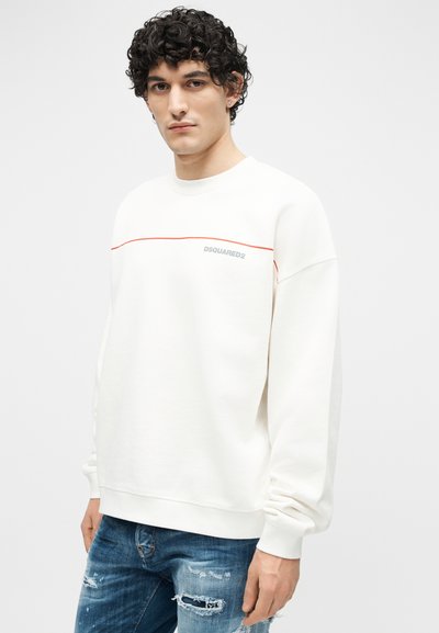 Νεαρός άνδρας με σγουρά μαλλιά, φοράει λευκό φούτερ DSQUARED2 με κόκκινη ρίγα και σκισμένο μπλε τζιν, σε απλό φόντο.