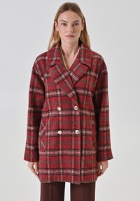 Donna che indossa un cappotto doppiopetto a quadri rossi e grigi con bottoni argentati, in posa su uno sfondo grigio chiaro uniforme.