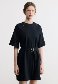 Νεαρή γυναίκα με ανοιχτόχρωμα μαλλιά φοράει μαύρο φόρεμα με κοντά μανίκια και ζώνη στη μέση, στέκεται μπροστά σε λευκό, μονόχρωμο φόντο.