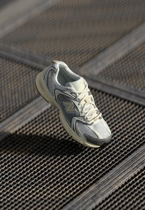 Weißer und grauer New Balance Sneaker schwebt über einer strukturierten Metallgitterfläche mit diagonalem Rautenmuster.