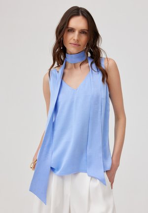 Kobieta o kręconych brązowych włosach, ubrana w jasnoniebieski top bez rękawów z dopasowaną apaszką na szyję oraz białe spodnie o szerokich nogawkach, stojąca z jedną ręką w kieszeni.