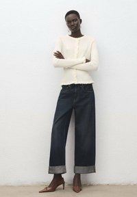 Cardigan bianco a coste in maglia con dettagli a bottoni, abbinato a pantaloni larghi in denim scuro con orli risvoltati e tacchi color cuoio con punta a punta.