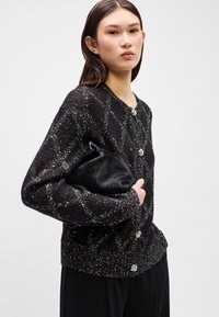 Black, textured cardigan with a sparkly pattern and silver buttons; model holds a small black clutch purse, displayed against a neutral background.
