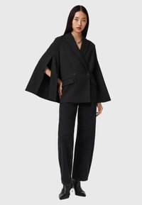 Femme portant un blazer cape noir avec un pantalon large noir et des bottes à talons noires, se tenant contre un fond gris clair uni.