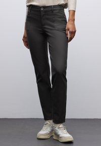 Osoba w czarnych dopasowanych spodniach i białych trampkach stoi na jednolitym jasnoszarym tle.