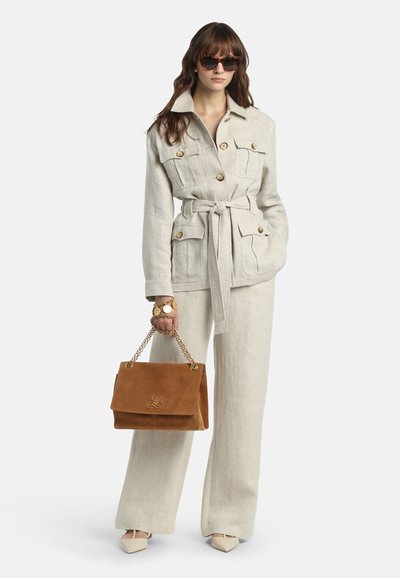 Femme portant une veste beige ceinturée et un pantalon, des lunettes de soleil écaille de tortue, tenant un sac à main en daim marron avec une chaîne dorée et un logo, chaussures beiges pointues.