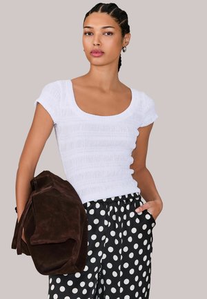 Femeie cu părul împletit, purtând un top alb cu textură, pantaloni negri cu buline albe, ținând o geantă mare maro din piele întoarsă.