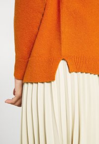 Orange Wolle Pullover mit gerippten Bündchen und einem gespaltenen Saum. In der unteren Bildhälfte ist ein plissierter cremefarbener Rock sichtbar.