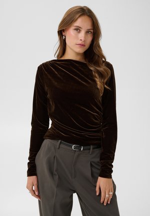 Jeune femme aux longs cheveux ondulés portant un haut en velours brun foncé à manches longues et un pantalon gris taille haute avec une ceinture noire.