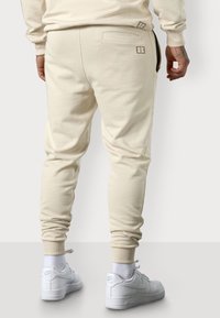Beige Jogginghosen aus weichem Stoff, mit schmalem Schnitt, zwei Gesäßtaschen und gerippten Bündchen, kombiniert mit weißen Sneakers.