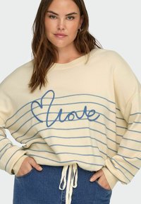 Femme portant un pull crème avec des rayures horizontales bleues, un cœur en corde bleue et le mot « amour », les mains dans les poches d'un jean en denim bleu.