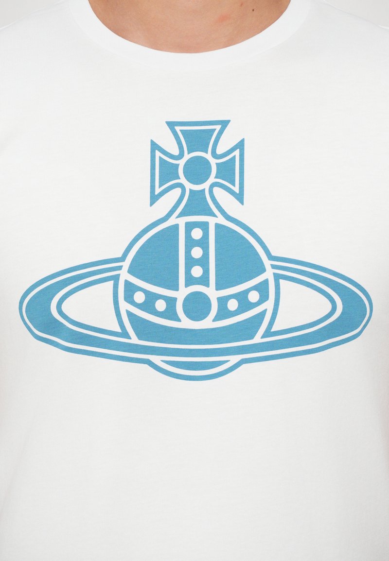 T-shirt blanc avec un graphique bleu représentant un globe entouré d'anneaux et surmonté d'une croix, mettant en valeur des designs géométriques et des accents circulaires.