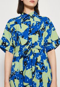 Košilové šaty v modré, zelené a černé vzorované látce. Mají krátké rukávy, límeček a pás na odstřihnutí v pase. Hladká, lehká textura.