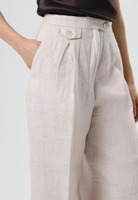 Lichtbeige linnenbroek met een hoge taille, plooien en zijzakken; afgewerkt met twee knopen aan de taille voor sluiting.