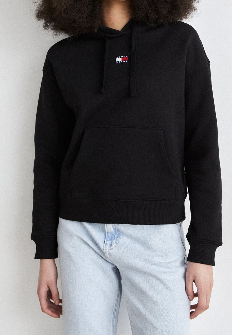 Sweat à capuche noir en tissu mélange de coton, avec une poche avant, des poignets côtelés, et un petit logo coloré sur la poitrine. Associé à un jean clair.