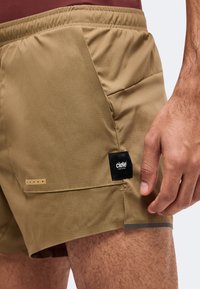 Gros plan d'une personne portant un short de sport beige avec une poche latérale, un logo noir Ciele Athletics et une bande réfléchissante près de l'ourlet.