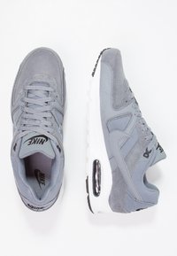 Grå Nike-sneakers med en blandning av mocka och mesh-material, med en vit mellansula, svarta accenter och synlig luftdämpning i hälen.