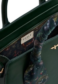 Lesklá kožená kabelka v lesnej zelenej farbe s podšívkou z patterned látky, obsahujúca značkovú nálepku. Rukoväť je obalená zladeným šálom.