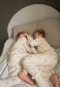 Kaksi lasta kermanvärisissä, pehmeissä pyjamasissa, joissa on hienovaraisia kukkakuvioita, kiedottuna toisiinsa valkoisella vuoteella, jossa on teksturoitu peitto.
